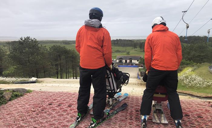 Two sit ski instructors in orange jackets each holding a sit ski are stood on top of an artificial ski slope preparing to ski.